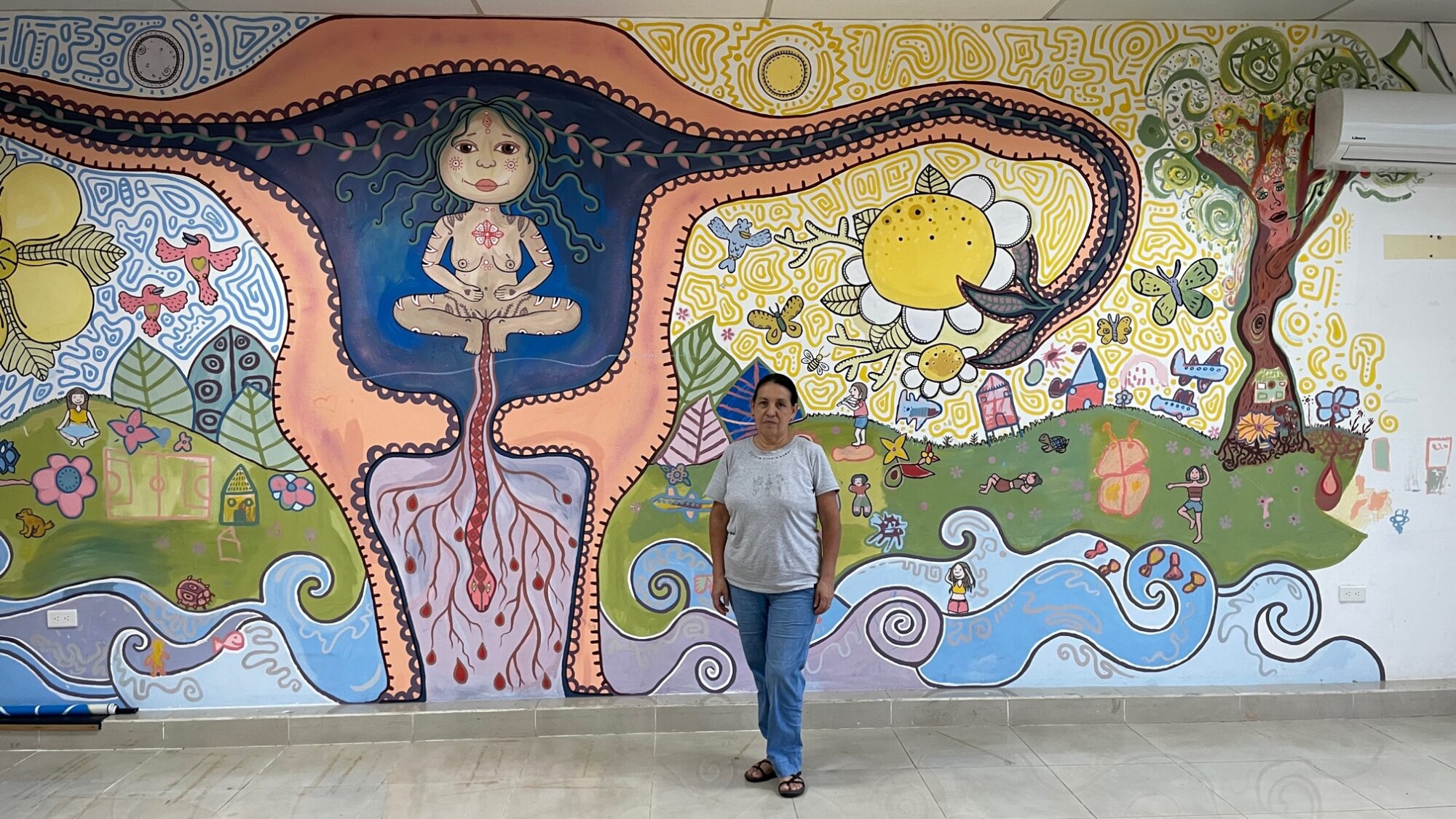 Inez, steht vor einer Wand in Casa Paula. An der Wand ist eine kunstvolle Abbildung eines Uterus zu sehen, in dem sich eine nackte Frau befindet. Rundherum sind Naturbilder, Blumen und Bäume aufgemalt.