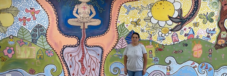 Inez, steht vor einer Wand in Casa Paula. An der Wand ist eine kunstvolle Abbildung eines Uterus zu sehen, in dem sich eine nackte Frau befindet. Rundherum sind Naturbilder, Blumen und Bäume aufgemalt.